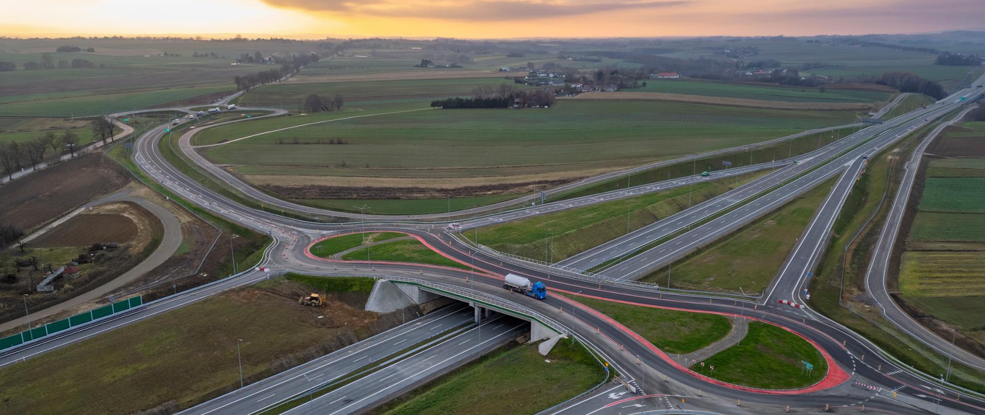 Widok z lotu ptaka na węzeł drogowy typu bezkolizyjnego: dwujezdniowa droga z pasem rozdziału, wiaduktem nad drogą główną, rondami turbinowymi oraz łącznicami wjazdowymi i zjazdowymi; otoczenie stanowią tereny rolnicze i rozproszona zabudowa, widok zachodzącego słońca.