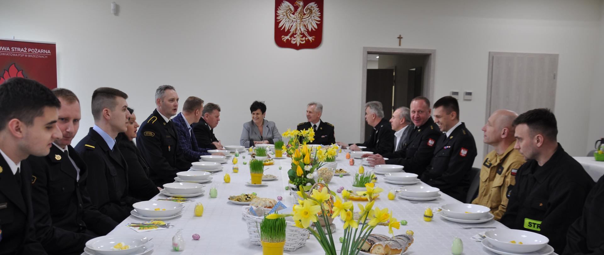 pomieszczenie w komendzie powiatowej psp w brzezinach przy zastawionym jedzeniem stole siedzą strażacy w umundurowaniu galowym oraz zaproszenie goście między innymi starościna Renata Kobiera, przewodniczący Rady Powiatu w Brzezinach Krzysztof Bogdański, kapelan powiatowy strażaków ks. Władysław Moczarski, prezes zarządu Oddziału Powiatowego ZOSP RP w Brzezinach dh Leszek Jarząb oraz były Komendant Powiatowy PSP w Brzezinach st. bryg. w st. sp. Dariusz Guzek