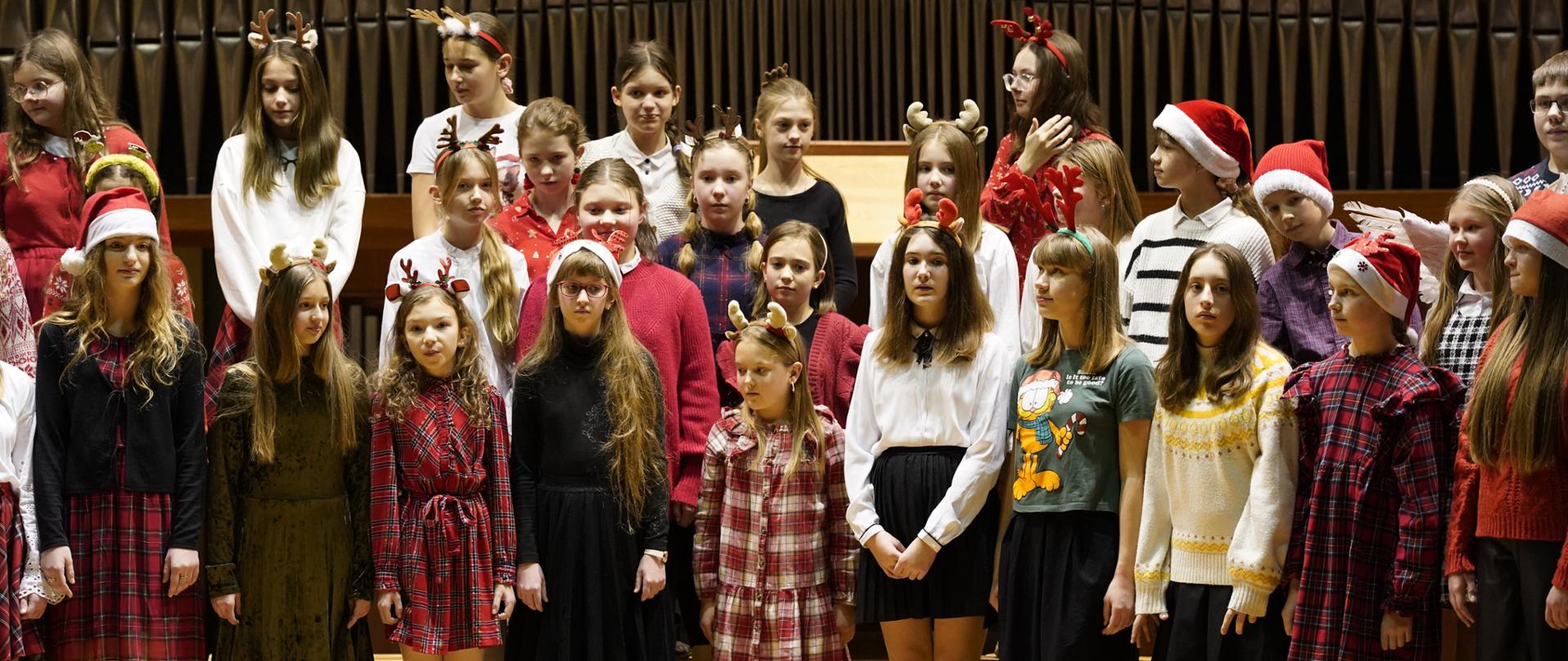 Chór dziecięcy na scenie sali koncertowej. Uczniowie stoją w trzech rzędach. Uczniowie ubrani w stroje bożonarodzeniowe. Niektóre z nich maja na głowie czapki Mikołaja lub rogi renifera. Dzieci stoją na drewnianych ławkach. Za nimi w tle organy.