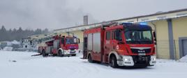 Dwa pojazdy ciężarowe straży pożarnej koloru czerwonego. Po prawej średni samochód ratowniczo-gaśniczy Jednostki Ratowniczo-Gaśniczej nr 1 w Bydgoszczy, po lewej samochód specjalny drabina mechaniczna z JRG1 w Bydgoszczy. W tle boczna elewacja hali koloru żółtego, w oddali zadymienie unoszące się nad halą.