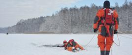 Na zdjęciu widać jezioro, w jeziorze jest przerębel w tle las. Widać trzech mężczyzn ubranych w ubrania niezatapialne , jeden z nich jest w wodzie, drugi leży na zamarzniętym lodzie i próbuje go wyciągnąć, trzeci stoi i trzyma linę asekuracyjną.