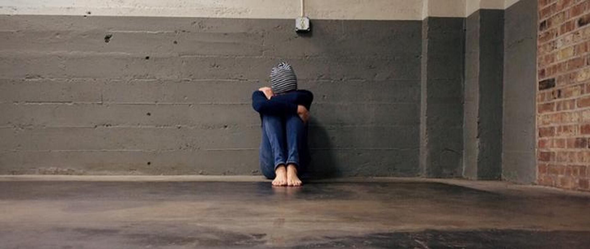 Kobieta bezdomna siedząca pod betonową ścianą z ukrytą twarzą