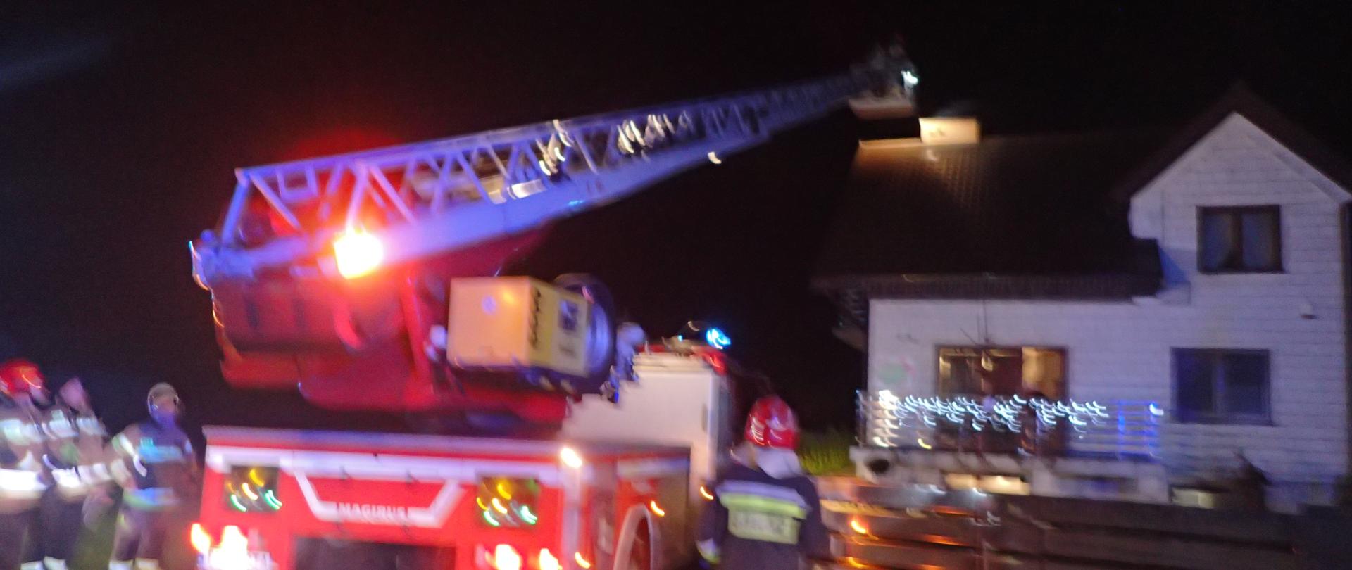Mechaniczna drabina pożarnicza sprawiana do dachu budynku mieszkalnego przy niej stoją strażacy, pora nocna