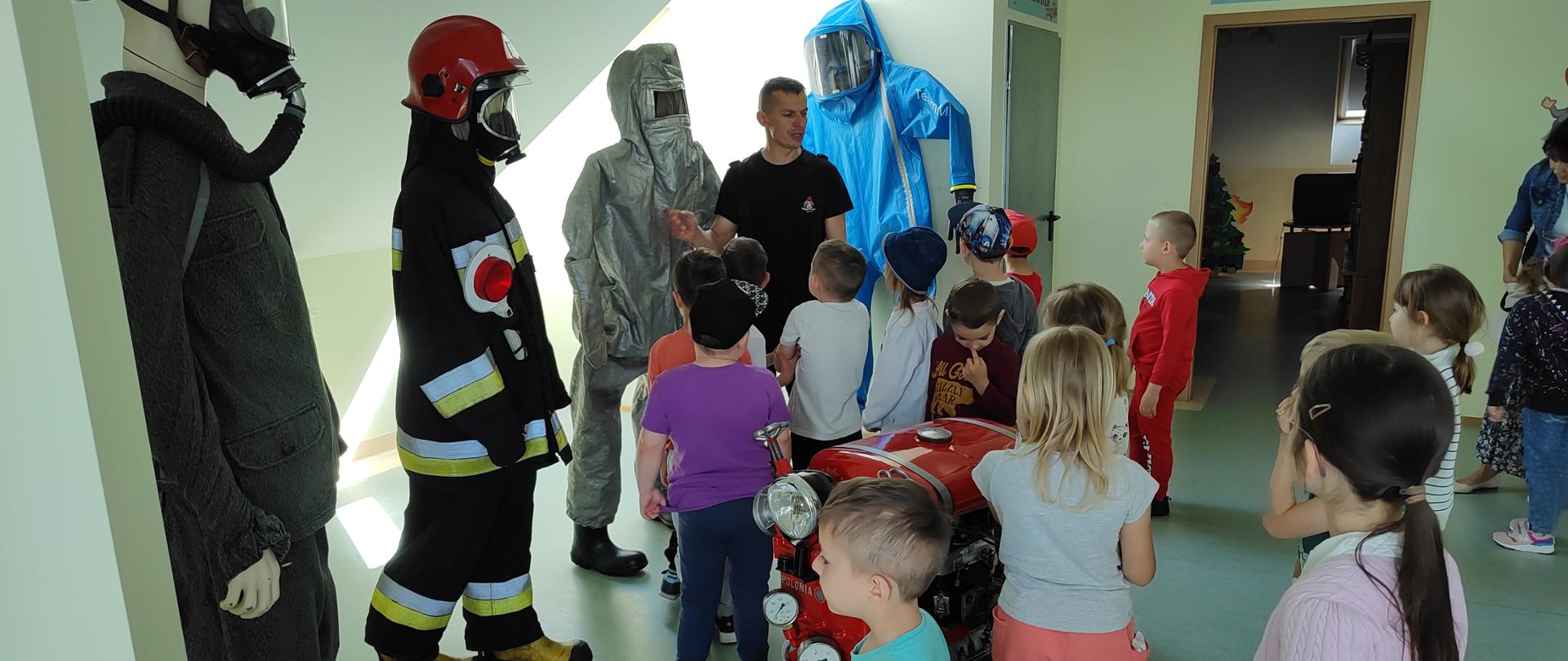 Dzieci stojące tyłem, strażak PSP na tle ubrań ochronnych w zależności od wykorzystania w działaniach. Po prawej stronie otwarte drzwi do pokoju i zamknięte srebrne drzwi do kotłowni. na pierwszym planie widać również motopompę pożarniczą. 