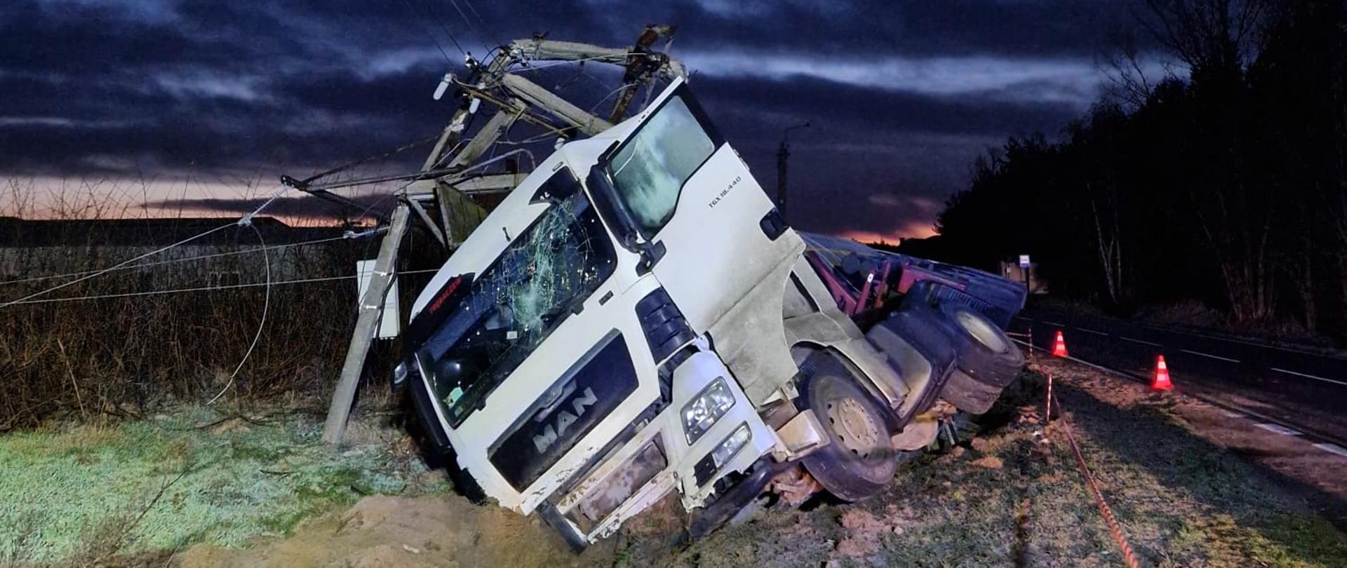 Na zdjęciu uszkodzony pojazd ciężarowy w rowie, przy drodze publicznej. Zdjęcie we wczesnych godzinach porannych. Oświetlenie sztuczne. 