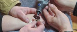 Kilka osób stoi wokół stołu, na którym leży plastikowy model dłoni ułożony na szarym ręczniku. Jedna osoba trzyma niewielkie narzędzie obrotowe z tarczą tnącą, skierowane w stronę modelu dłoni, jakby wykonywała precyzyjne cięcie lub pokaz demonstracyjny. Inna osoba stabilizuje model dłoni, trzymając go delikatnie od spodu. W tle widać dłonie pozostałych uczestników, którzy przyglądają się działaniu. Na stole stoi także plastikowa butelka z wodą. Cała scena wygląda na praktyczne ćwiczenie lub instruktaż w warunkach warsztatowych.