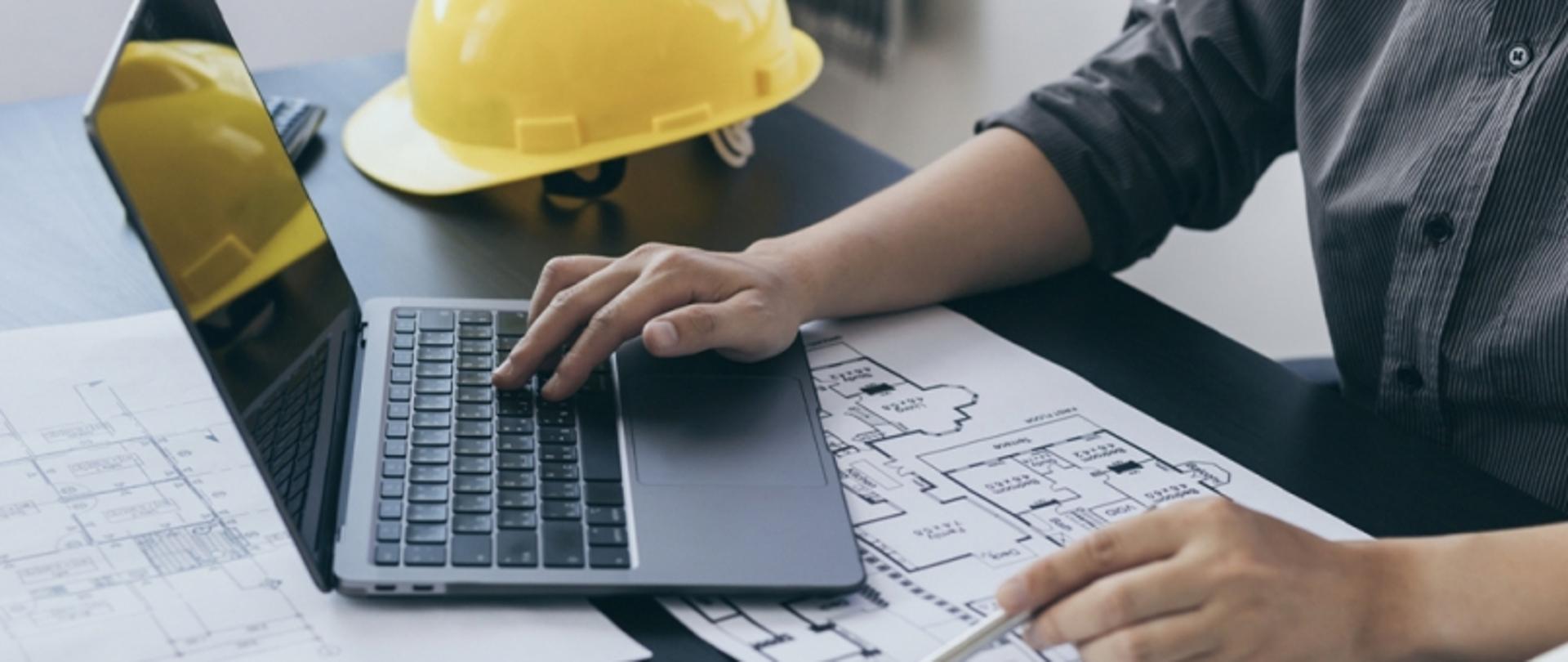 Osoba pracująca przy biurku z laptopem i planami architektonicznymi, z żółtym kaskiem ochronnym na blacie.