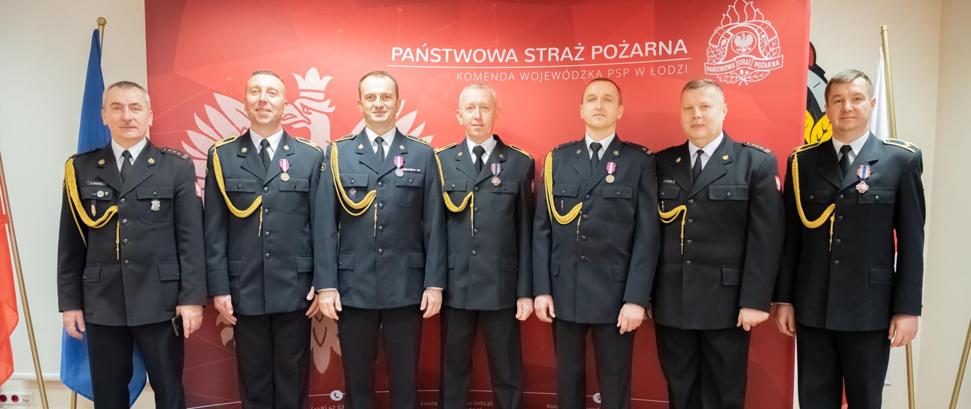 Strażacy ubrani w wyjściowe mundury stoją jeden obok drugiego. Pomieszczenie w którym się znajdują ma jasne ściany i podłogę. Za strażakami stoi czerwona ścianka z białym logiem Państwowej Straży Pożarnej oraz napisem Państwowa Straż Pożarna Komenda Wojewódzka PSP w Łodzi. Z lewej i prawej strony strażaków stoją flagi.