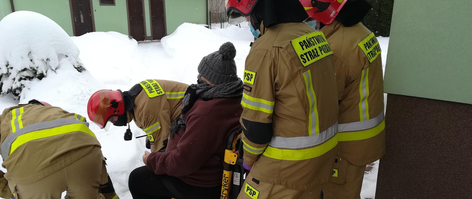 Na zdjęciu strażacy w ubraniu specjalnym w kolorze piaskowym oraz w hełmach, znoszą starszą kobietę na wózku transportowym. W tle zabudowania oraz dużo śniegu.