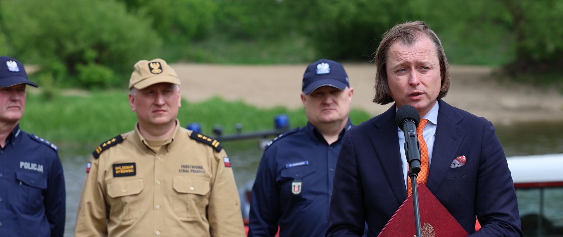 Briefing prasowy na plaży w Pomiechówku. Do mikrofonu przemawia Wojewoda Mazowiecki Mariusz Frankowski. Za nim stoją Zastępca Mazowieckiego Komendanta Wojewódzkiego PSP st. bryg. Waldemar Wysowski oraz dwóch funkcjonariuszy Policji. 