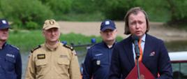 Briefing prasowy na plaży w Pomiechówku. Do mikrofonu przemawia Wojewoda Mazowiecki Mariusz Frankowski. Za nim stoją Zastępca Mazowieckiego Komendanta Wojewódzkiego PSP st. bryg. Waldemar Wysowski oraz dwóch funkcjonariuszy Policji. 