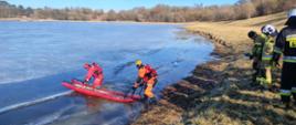 dwaj strażacy wchodzą do wody z czerwonymi saniami lodowymi. Do strażaków przyczepione są liny które trzymaja strażacy stojący na brzegu