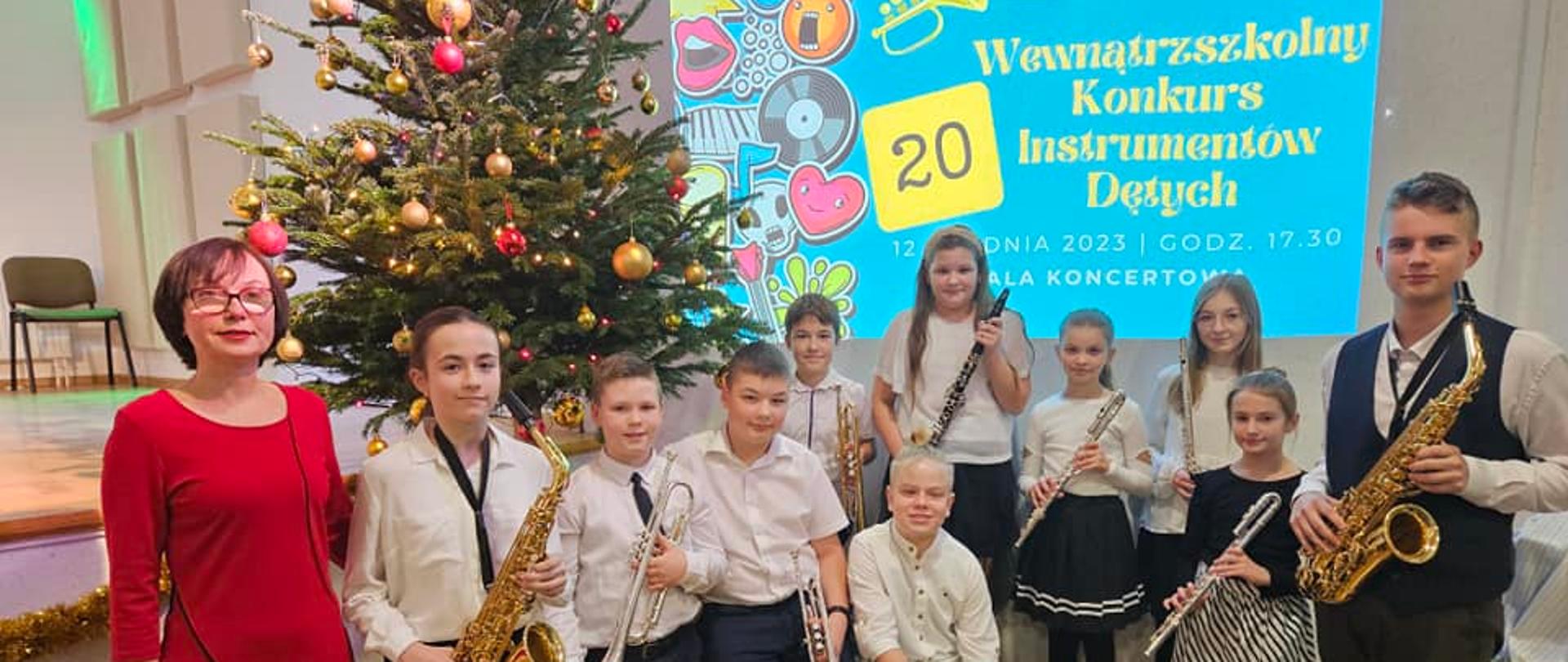 Uczniowie trzymający w rękach woje instrumenty przed szkolnym konkursem instrumentów dętych. W tle choinka oraz wyświetlony plakat konkursu.