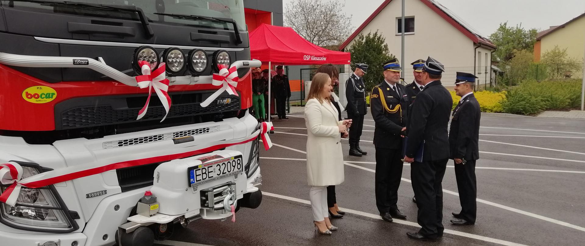 na zdjęciu widzimy komendantów i strażaków w ubraniach wyjściowych oraz zaproszonych gości podczas uroczystości Gminnych obchodów Dnia Strażaka z lewej strony samochód straży przepasany wstęgą biało-czerwoną w tle budynki