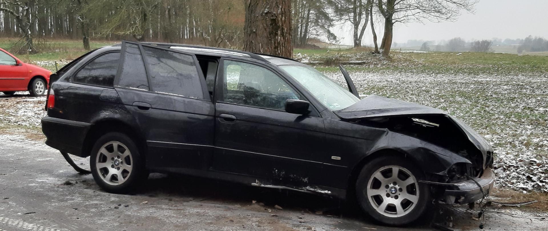 Czarny samochód marki BMW stoi na drodze z rozbitym przodem i otwartymi tylnymi drzwiami. Za pojazdem widać osobowy czerwony samochód. 