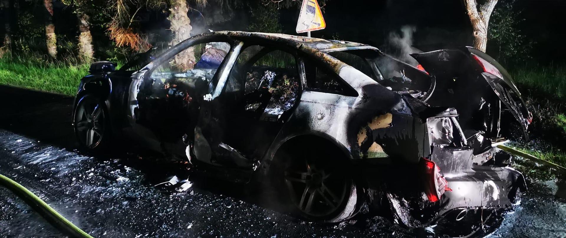 Zdjęcie przedstawia spalony wrak samochodu na drodze podczas pory nocnej.