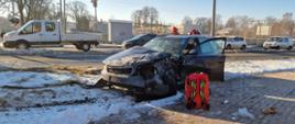 Zdjęcie przedstawia miejsce wypadku drogowego w warunkach zimowych. Na pierwszym planie widoczny jest poważnie uszkodzony, ciemnoszary samochód osobowy marki Audi (prawdopodobnie model A4 lub A6 kombi), który zjechał z jezdni na pobocze i pas zieleni. Na miejscu pracują już służby ratunkowe.