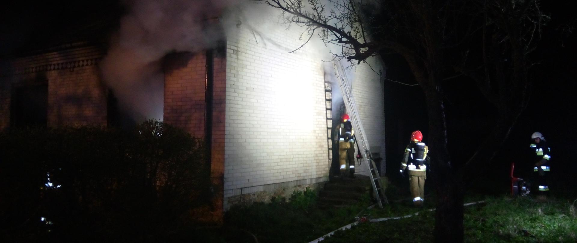 Zdjęcie przedstawia 3 strażaków przed budynkiem z którego wydobywa się dym