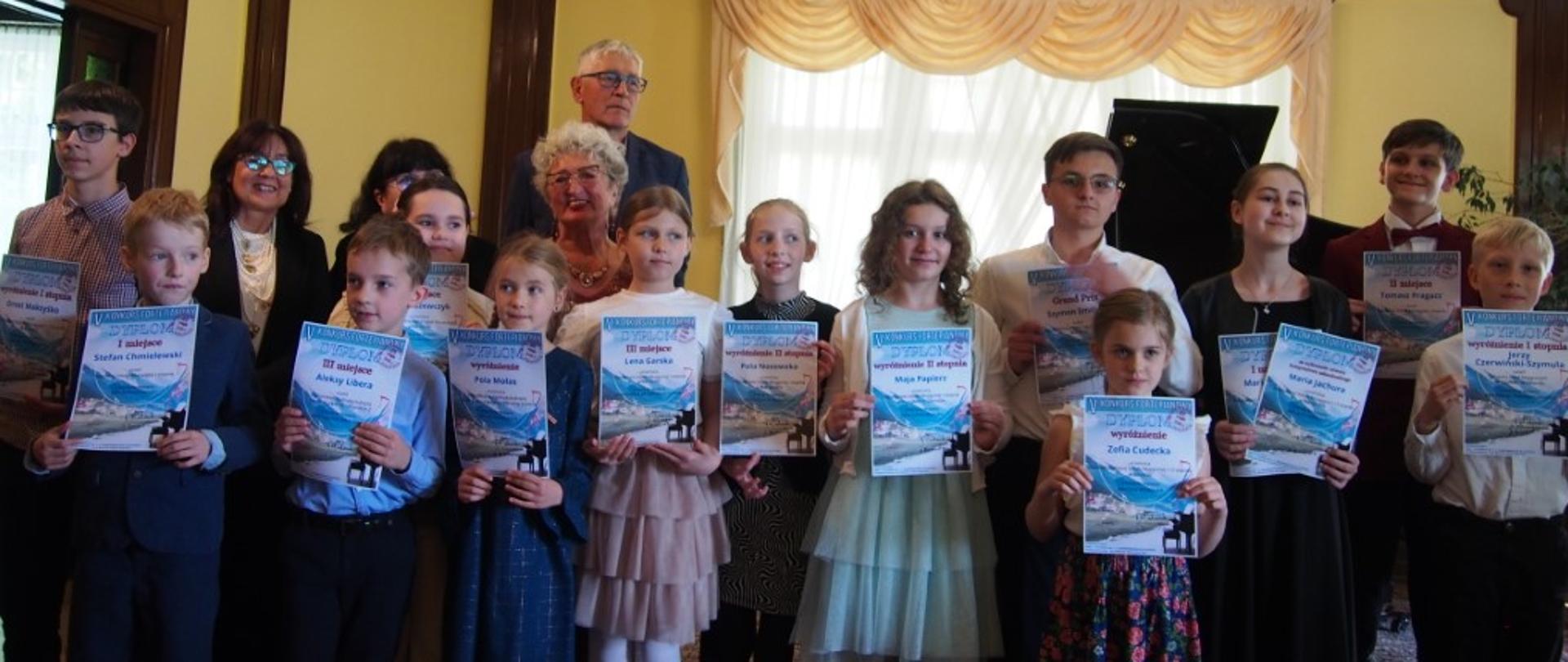 Zdjęcie kolorowe. Grupa młodych osób, uczestników konkursu, trzymających dyplomy lub świadectwa. Stoją w rzędzie w eleganckim wnętrzu z drewnianą boazerią i żółtymi ścianami. W tle żyrandol i okna z zasłonami.