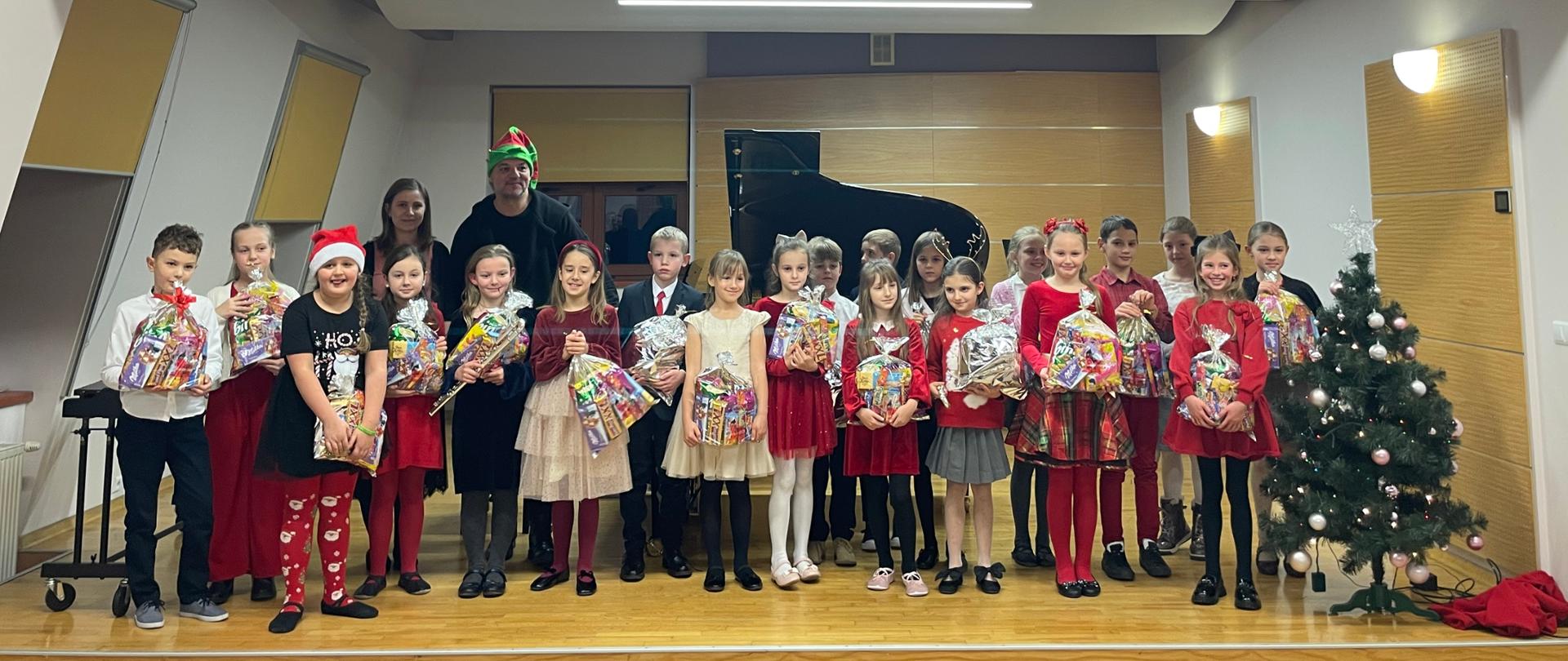 Na zdjęciu widać grupę około dwudziestu kilku dzieci stojących razem na scenie w jasnym, nowoczesnym pomieszczeniu. Większość dzieci jest odświętnie ubrana — dziewczynki w eleganckich sukienkach, często w kolorach czerwieni lub beżu, a chłopcy w koszulach i marynarkach. Wszyscy trzymają w rękach kolorowe paczki upominkowe owinięte w przezroczyste folie.
Pośrodku stoi dorosły mężczyzna w czarnej bluzie, na głowie ma zielono-czerwoną czapkę elfa, więc wygląda jak pomocnik świętego Mikołaja. W tle znajduje się duży, czarny fortepian. Z prawej strony stoi mała choinka ozdobiona bombkami. Cała grupa uśmiecha się do zdjęcia, tworząc ciepłą, świąteczną atmosferę.