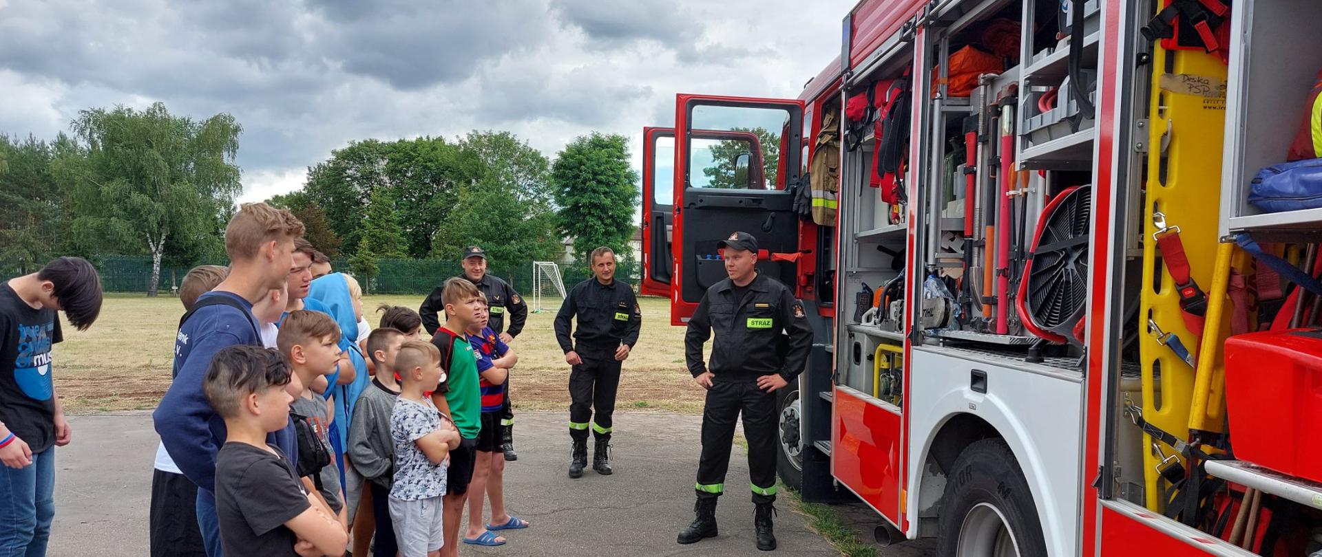 Na zdjęciu widać dzieci stojących z boku samochodu ratowniczo-gaśniczego w szeregu, którzy słuchają trzech strażaków stojących przed samochodem strażackim. W tle widać boisko piłkarskie