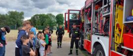 Na zdjęciu widać dzieci stojących z boku samochodu ratowniczo-gaśniczego w szeregu, którzy słuchają trzech strażaków stojących przed samochodem strażackim. W tle widać boisko piłkarskie