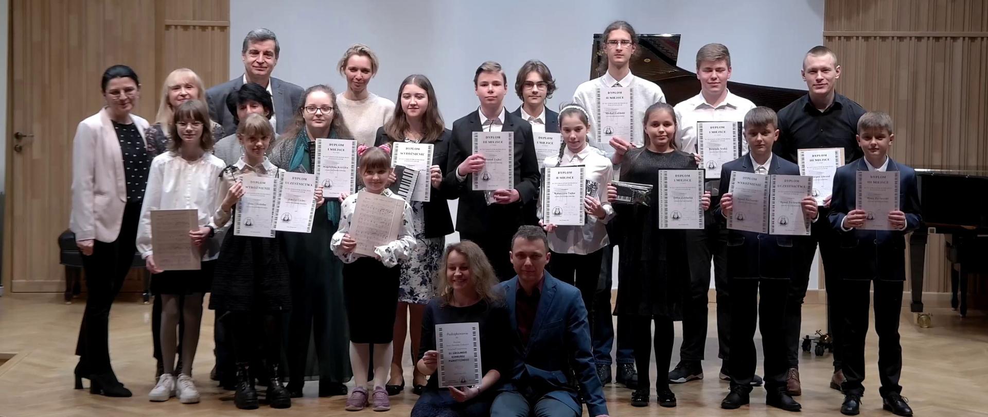 Zdjęcie: na estradzie szli koncertowe stoją uczestnicy oraz członkowie Jury i organizatorzy konkursu szkolnego. Laureaci trzymają w rękach dyplomy. W tle na pofalowanej ścianie wyświetlony jest napis XII Szkolny Konkurs Pianistyczny 30 marca 2023.