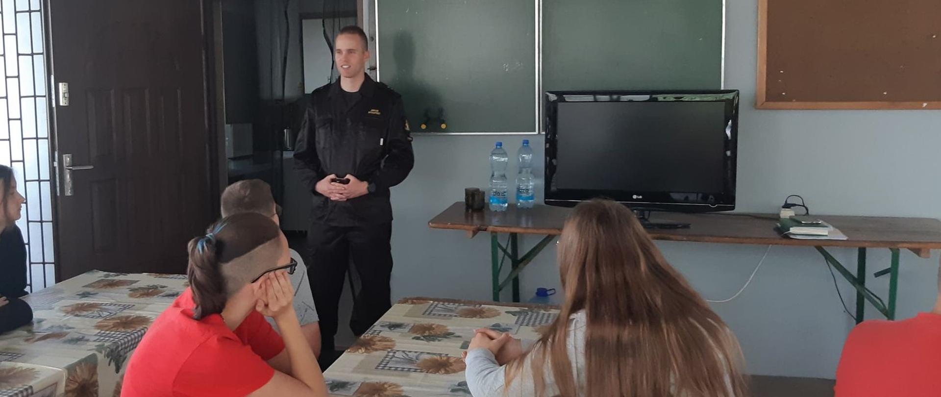 Zdjęcie przedstawia strażaka przeprowadzającego edukację dla dzieci i młodzieży na wakacyjnym obozie strażackim.
W tle świetlica.
