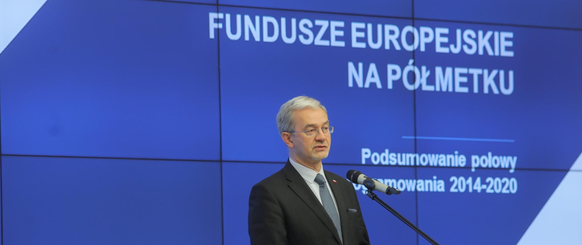 Na tle niebieskiego ekranu z napisem "Fundusze Europejskie na półmetku" stoi minister Jerzy Kwieciński, który przemawia do mikrofonu