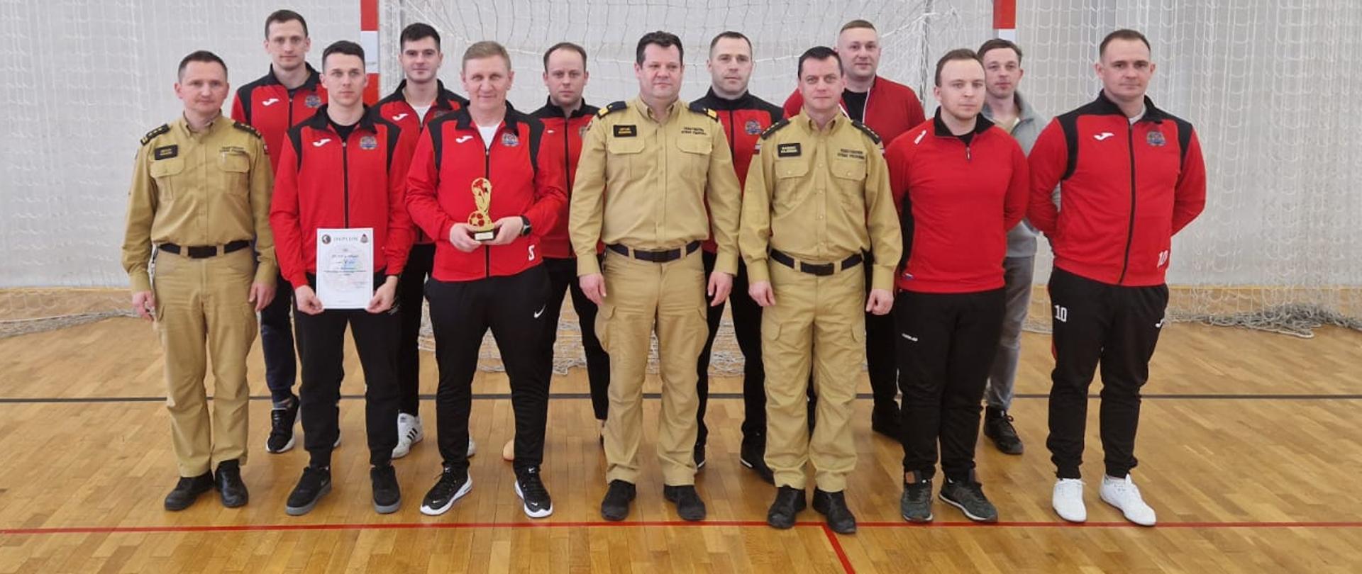 Zdjęcie przedstawia reprezentację drużyny Komendy Powiatowej Państwowej w Mławie wraz z przedstawicielem Komendy Wojewódzkiej Państwowej Straży Pożarnej, Komendantem Wojewódzkim nadbrygadierem Arturem Gonerą, st. bryg. Artur Wójcik naczelnik wydziału operacyjnego Komendy Wojewódzkiej PSP oraz dowódca jednostki JRG Mława oraz. Osoby znajdują się w sali sali gimnastycznej szkoły.