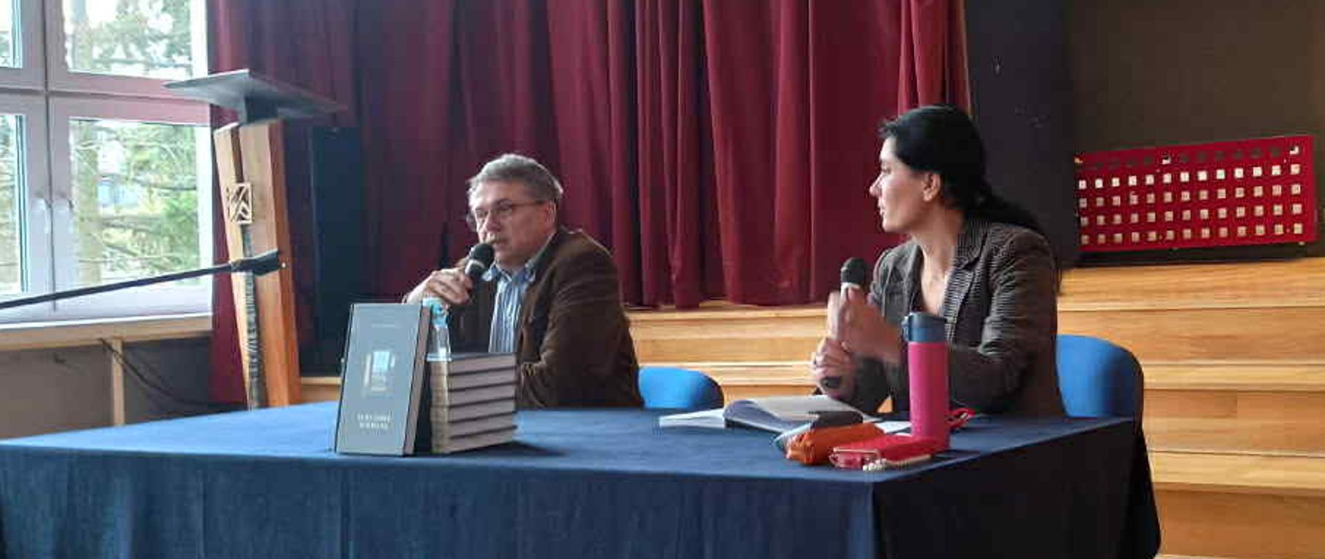 Mężczyzna i kobieta siedzą za stołem przykrytym niebieskim obrusem w dużym pomieszczeniu. Po lewej stronie duże okno (za oknem jasno, widać zieleń drzew). Na sali czerwone kotary, scena z czerwonymi kurtynami, nad sceną widać fragment godła Polski. Po prawej stronie, w głębi sceny, duży obraz mężczyzny z siwymi włosami i brodą.