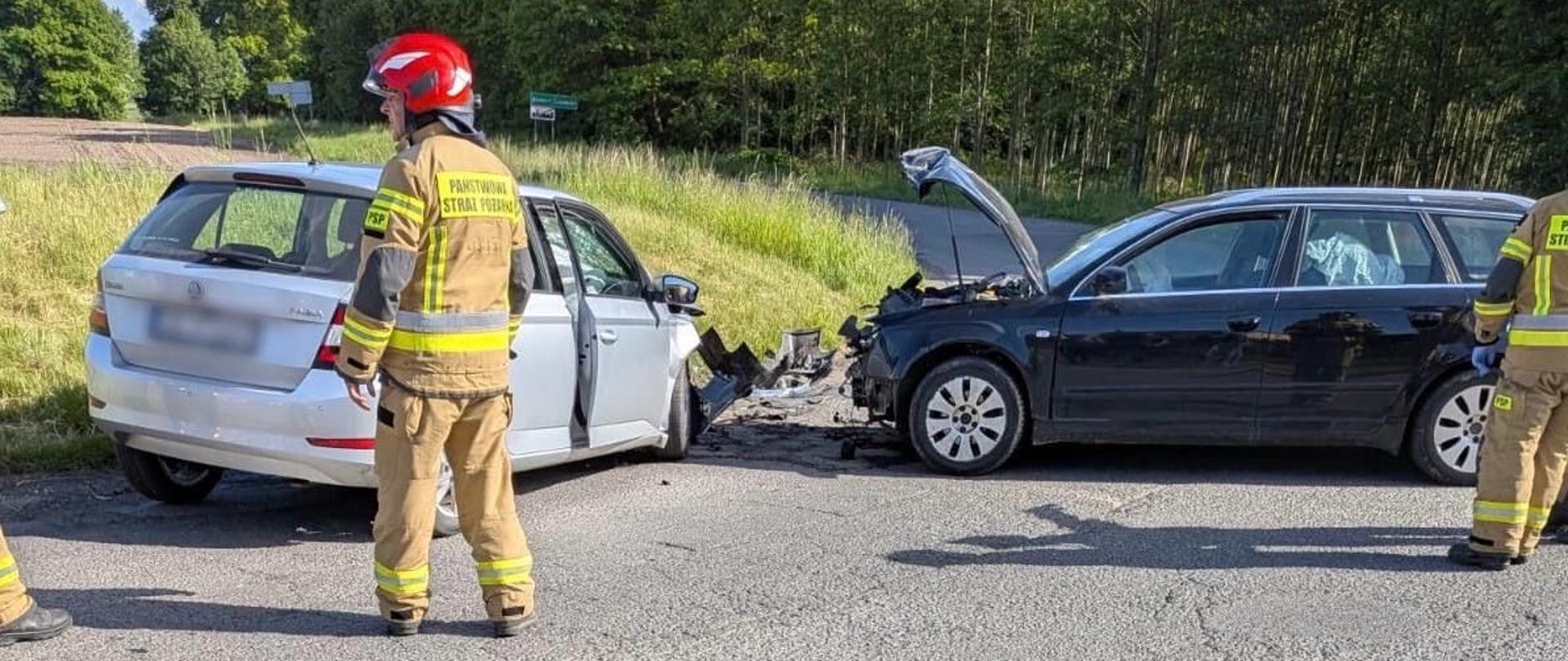 Na zdjęciu znajdują się dwa wraki samochodów oraz strażak w ubraniu specjalnym, w tle widać drzewa