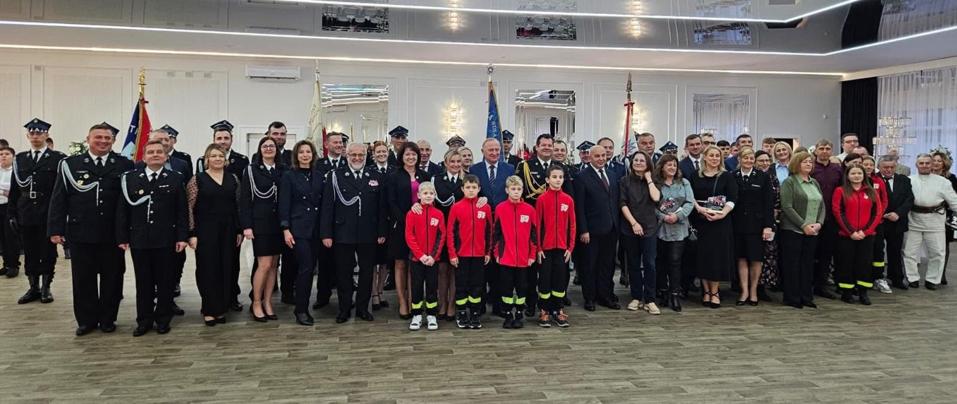 Uczestnicy wydarzenia pozują do zdjęcia na parkiecie białej jasnej sali z lustrami na ścianach.
