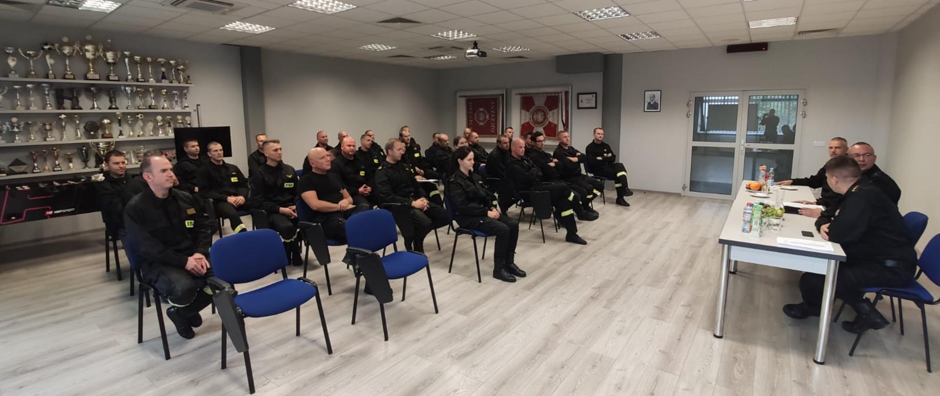 Zdjęcie wykonano na świetlicy KP PSP w Wolsztynie. Na zdjęciu widoczna komisja egzaminacyjna siedząca przy stole oraz funkcjonariusze przystępujący do egzaminu, siedzący na krzesłach.