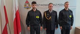 Komendant wraz ze dwoma strażakami po bokach na tle loga PSP, obok flagi państwowe