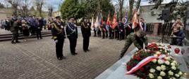Trzech przedstawicieli służb mundurowych stoi przed pomnikiem, żołnierz składa wieniec. Za nimi stoją uczestnicy oraz poczty sztandarowe.