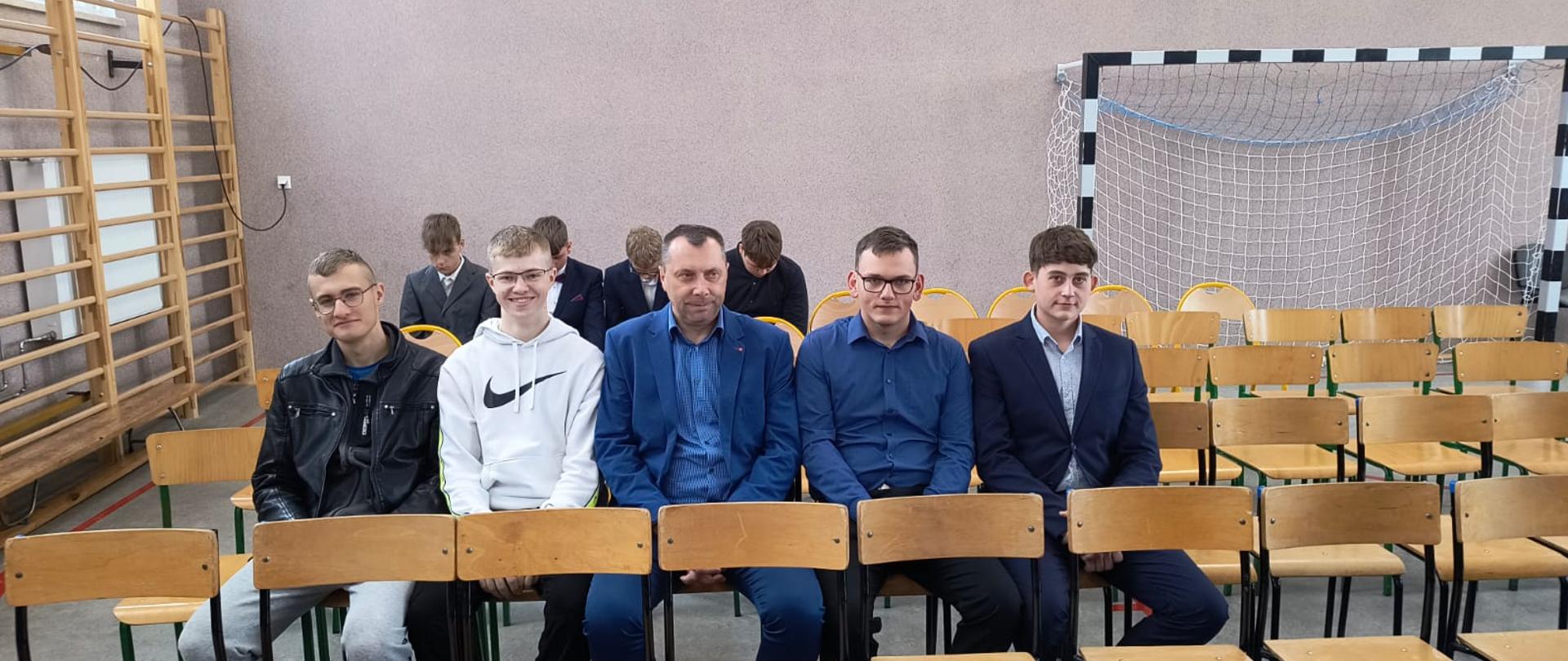 Uczniowie i nauczyciel siedzą w sali gimnastycznej przed konkursem.