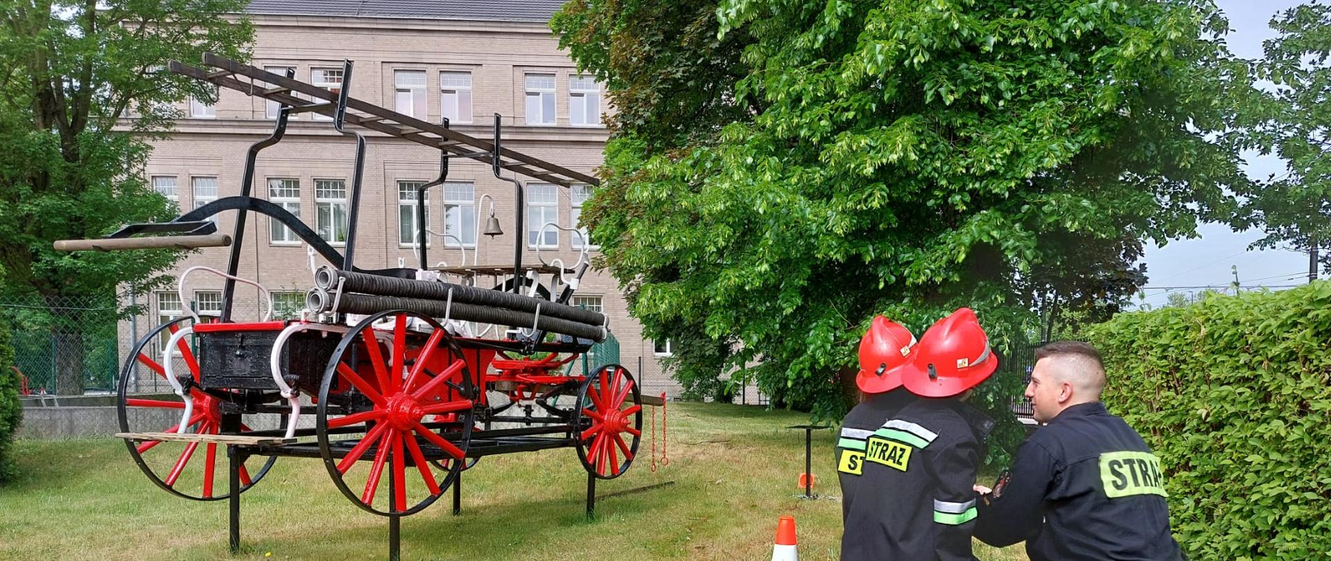 Dzieci w towarzystwie kucającego strażaka, ubrane w hełmy i kurtki strażackie trzymają prądownicę wraz z linią wężową, z prawej strony znajduje się sikawka konna.