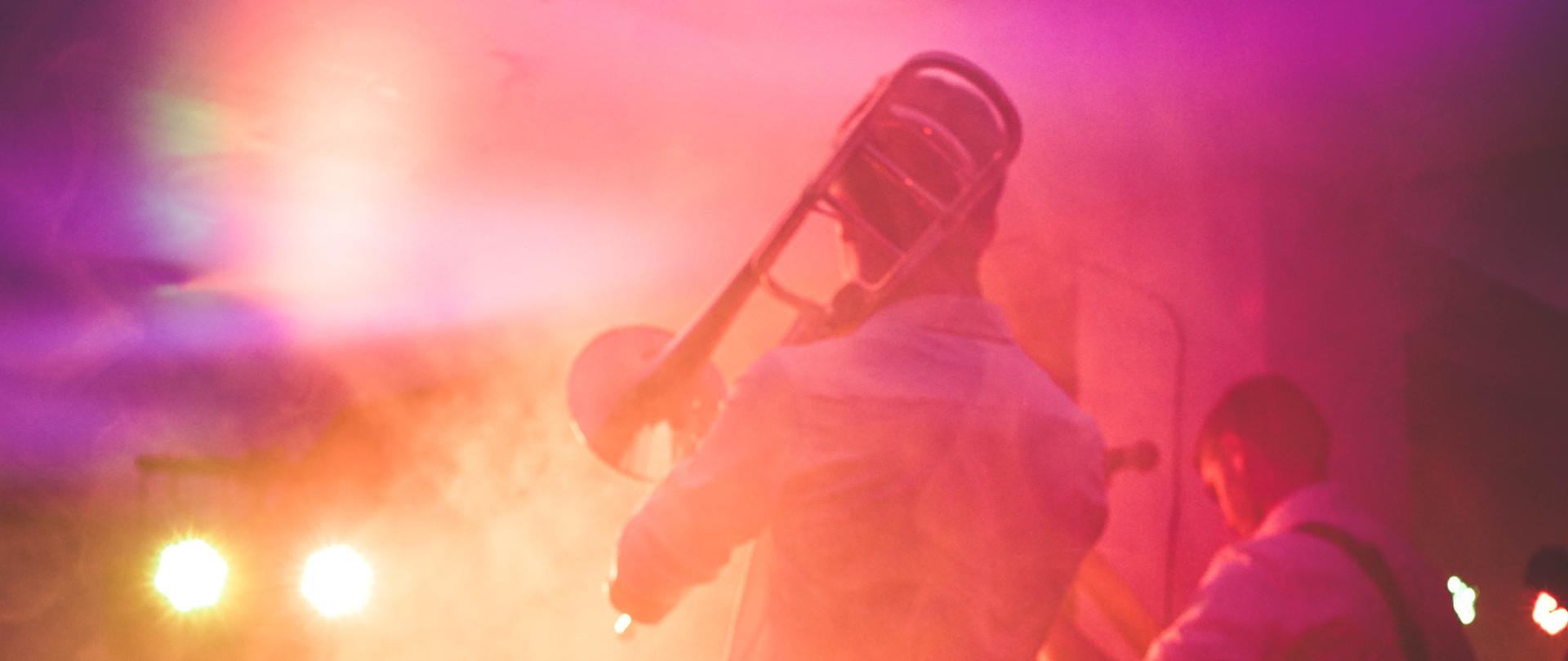 Zdjęcie przedstawia dwie osoby stojące na scenie plecami do obserwatora. Osoba po lewej stronie gra na puzonie, osoba po prawej gra na gitarze.