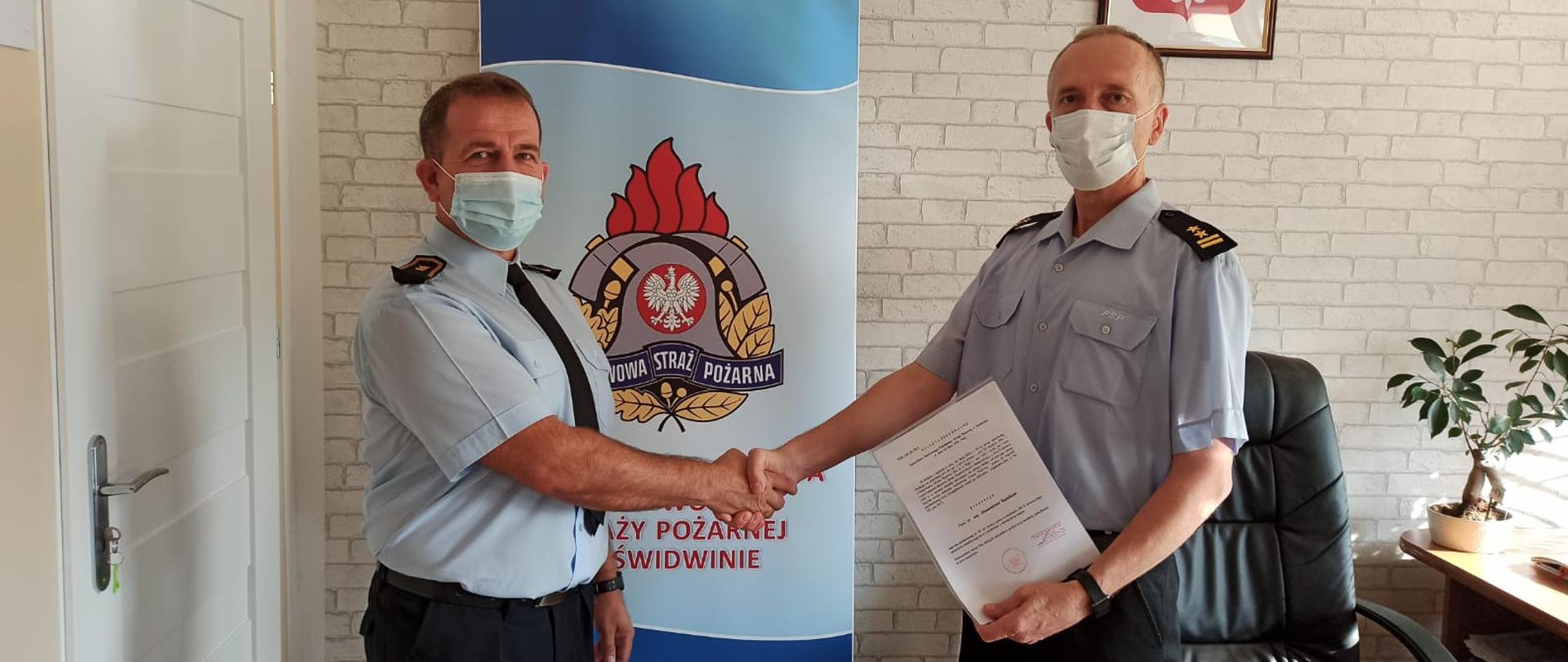 Zdjęcie przedstawia dwóch mężczyzn ubranych w mundury służbowe oraz maseczki chirurgiczne, jeden z nich trzyma w rękach dokumenty. Za nimi widnieje baner z logiem PSP oraz napisem: "Komenda Powiatowa Państwowej Straży Pożarnej w Świdwinie". Za banerem widzimy ścianę z tapetą w szare cegły. Na ścianie wisi Godło Rzeczypospolitej Polskiej. Zdjęcie wykonano w gabinecie Komendanta Powiatowego PSP w Świdwinie.