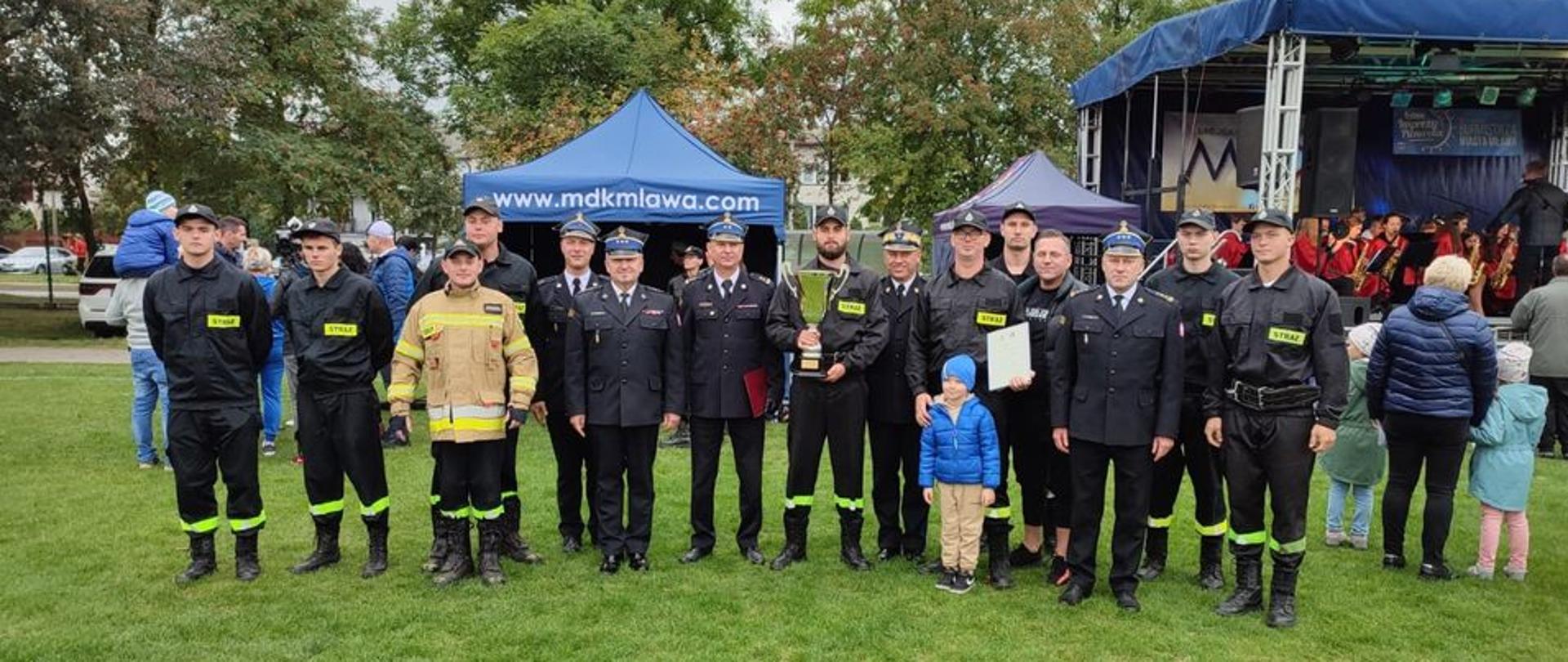 Strażacy stoją w szeregu. Jeden z nich trzyma puchar, drugi dyplom. W tle widać scenę oraz namiot z napisem "www.mdkmlawa.com.
