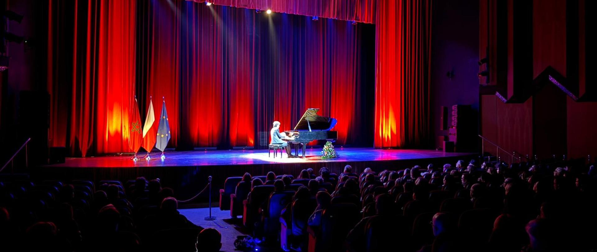 Na scenie w głębi pianista przy fortepianie, po lewej flagi Polski, Maroka i UE, pierwszoplanowo publiczność w pół cieniu