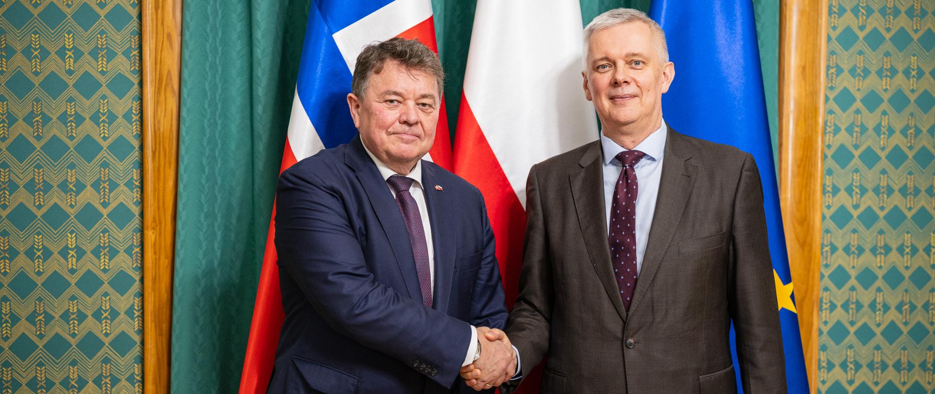 Po prawej stronie minister Tomasz Siemoniak, po lewej stronie ambasador Norwegii Øystein Bø. Panowie witają się na tle trzech flag - UE po prawej (niebieska z żółtymi gwiazdkami), flaga Polski w środku (biało-czerwona) oraz flaga Norwegii po lewej (czerwono-niebiesko-biała). W tle widać również zielone kotary, drewniane sztukaterie oraz zieloną tapetę w złote wzory.
