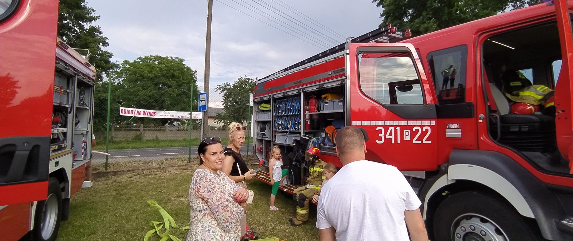 Na zdjęciu widzimy ludzi stojących przy samochodzie strażackim. Samochód koloru czerwonego, żaluzje podniesione do góry widzimy sprzęt ratowniczy jaki znajduje się na pojeździe. 