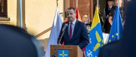 Wojewoda przemawia przy mównicy do strażaków za nim sztandary oraz inne osoby