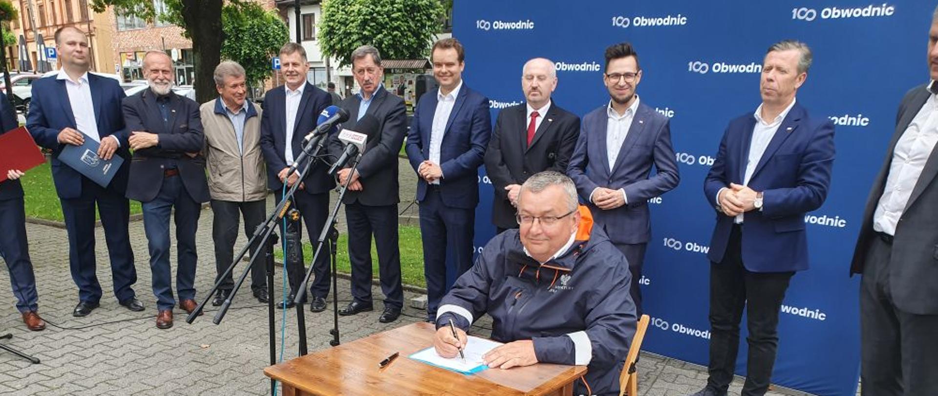 Na zdjęciu widać grupę ludzi stojących na granatowej ściance z napisem 100 obwodnic. Jeden z mężczyzn siedzi przy stoliku i podpisuje dokument. Wszyscy są uśmiechnięci i zadowoleni. 