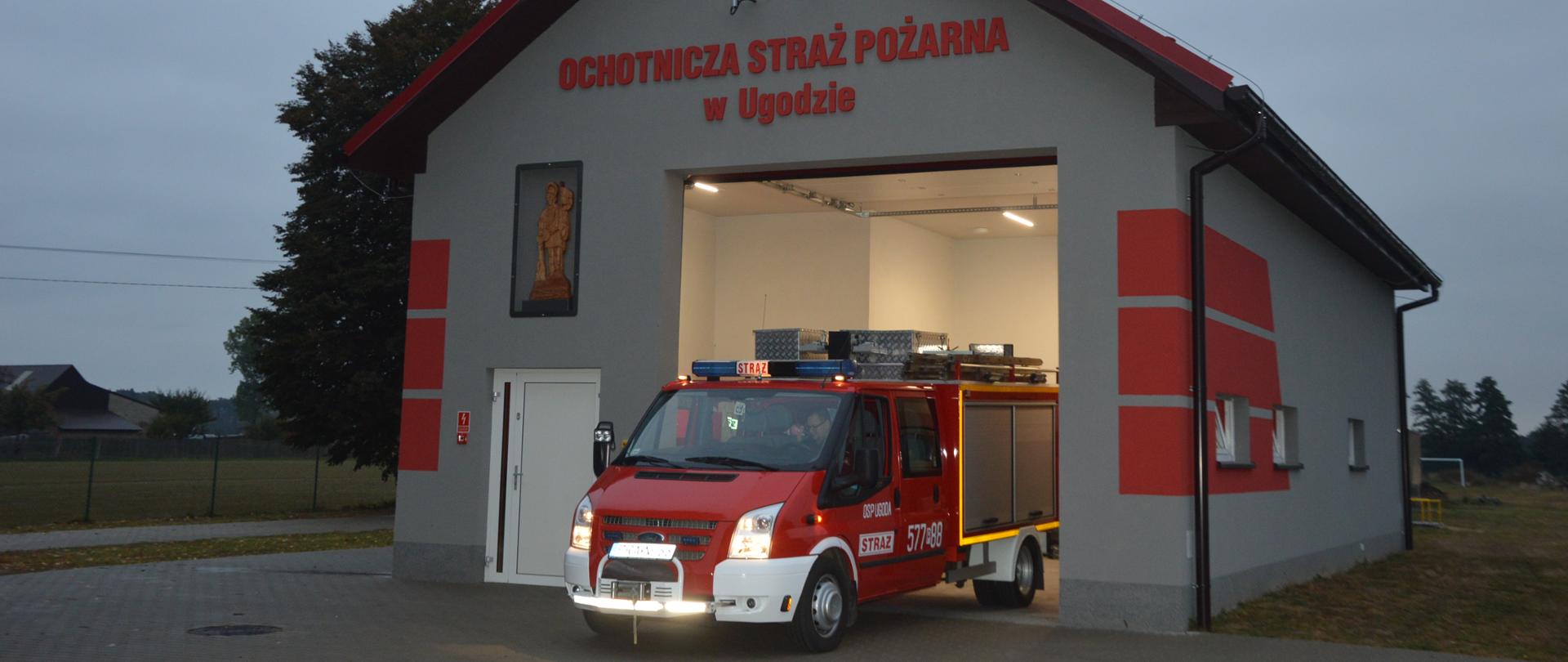 Budynek nowej remizy OSP Ugoda. W bramie ustawiony jest samochód pożarniczy. Z lewej strony na frontowej ścianie znajduje się figura św. Floriana. Na górze napis w kolorze czerwonym "Ochotnicza Straż Pożarna w Ugodzie". Nad napisem umieszczona syrena alarmowa. W tle drzewa.