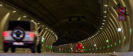 Wnętrze oświetlonego tunelu drogowego z jadącymi pojazdami; po lewej stronie widać rozpędzony samochód terenowy, a po prawej tablice zmiennej treści z ograniczeniem prędkości do 90 km/h i zakazem wyprzedzania.