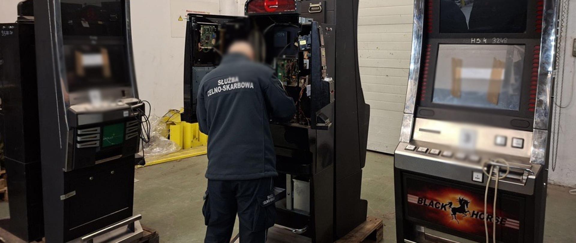 Funkcjonariusz Służby Celno-Skarbowej przy automacie do gier z otwartą obudową i sprawdza jego wnętrze; obok stoją inne urządzenia tego samego typu.