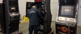 Funkcjonariusz Służby Celno-Skarbowej przy automacie do gier z otwartą obudową i sprawdza jego wnętrze; obok stoją inne urządzenia tego samego typu.